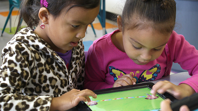 Two preschool girls play the Early Math with Gracie & Friends Treasure Bubbles app, each child with one finger moving characters on the iPad screen.