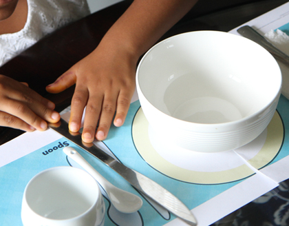 A young girl places a knife on a paper placement between a bowl and a spoon.