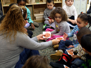A preschool teacher sitting on the floor hands paper cookie cut-outs to several children.