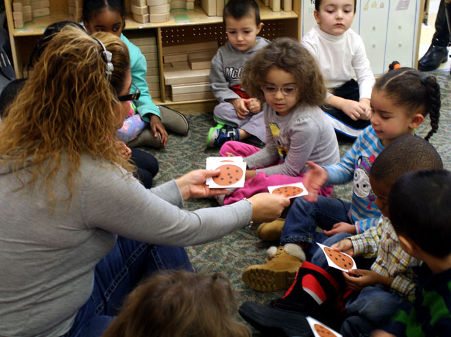 A preschool teacher sitting on the floor hands paper cookie cut-outs to several children.