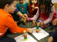 A group of preschoolers sit around two unequal towers of blocks. Two children point to a tower, counting its blocks.