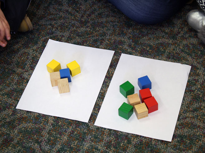 Colorful wooden blocks sit on two pieces of white paper in a group of five and a group of seven.