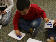 A boy in a red shirt sitting on the floor with his classmates puts blue dot stickers on a white piece of paper.
