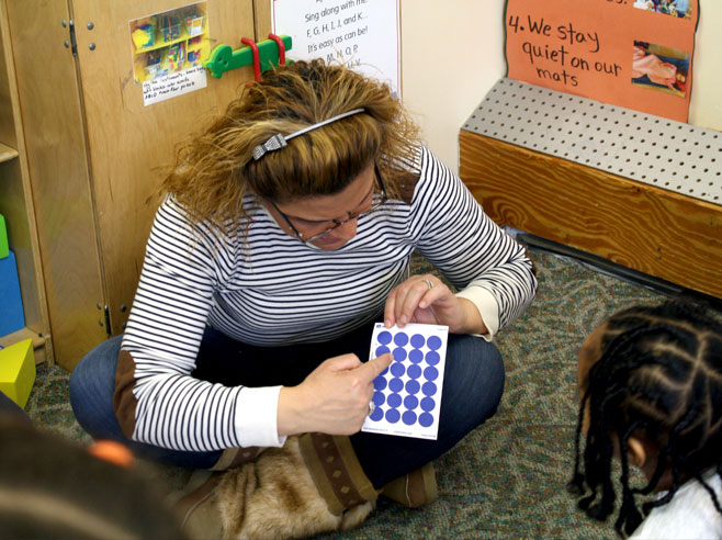 A preschool teacher sits on the floor in front of several children and points to a sheet blue dot stickers.