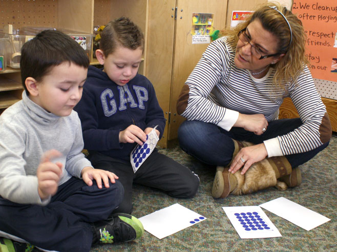 Two boys sit on the carpet. One peels a dot sticker from a sheet. The teachers sits nearby and talks with them.