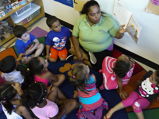 A teacher holds up the book as she reads. Young children sit around her and look on.