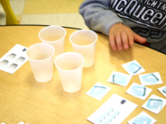 A preschool child in a gray sweatshirt reaches for the Drink Up Card Game materials on the table in front of him.