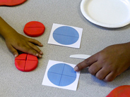 Close-up of a teacher’s hand and a child’s hand pointing to two red play dough circles cut to match blue paper templates.