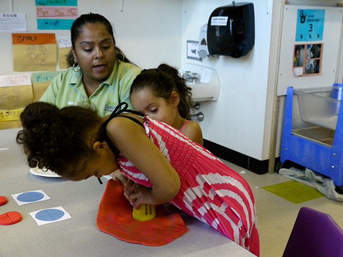 A young girl cuts red play dough into a circle while her teacher and a classmate look on.