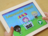 Close-up of a child’s hands holding an iPad with the homepage of the Gracie and Friends Lemonade Stand app on-screen.