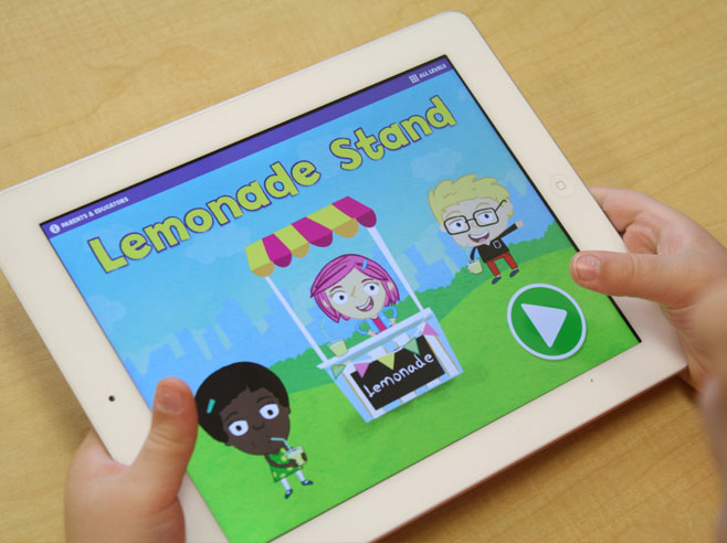 Close-up of a child’s hands holding an iPad with the homepage of the Gracie and Friends Lemonade Stand app on-screen.