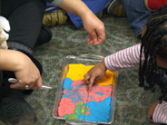 A girl in a striped shirt points to the center of a play dough lasagna that is on the carpet between her and her teacher.