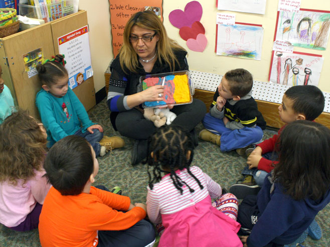 Preschool children sit on a carpet with their teacher, who holds a tray of pretend lasagna and a plastic knife across its center.