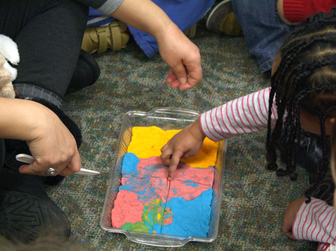 A girl in a striped shirt points to the center of a play dough lasagna that is on the carpet between her and her teacher.