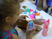 A girl puts a small plastic bottle into a brown paper bag. Other small toys are on the table in front of her.