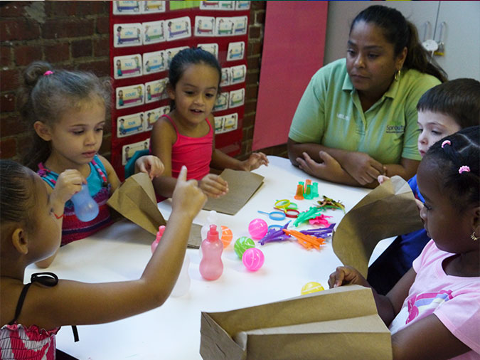 Five children and their teacher hold paper bags and look at toys on the table.