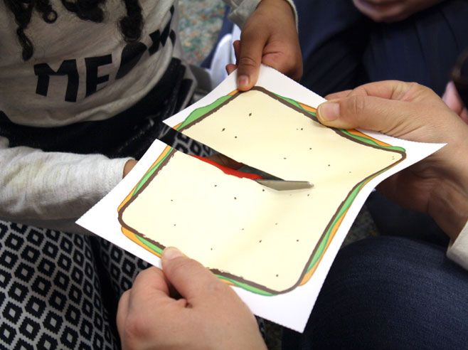 Close-up of a child cutting a paper sandwich held by a teacher.