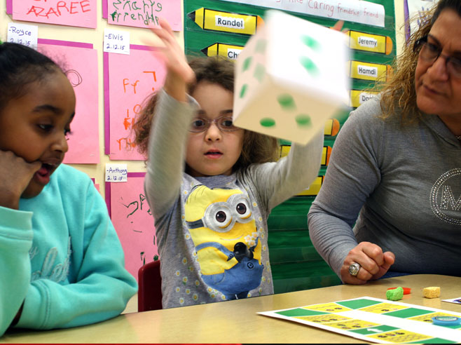 A girl throws a large die into the air over the Park Play Board Game while a classmate and teacher look on.