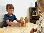 A boy and a girl position two different block towers in relation to each other based on a paper card on a table.