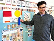 A man stands in front of a preschool bulletin board and places a blue rectangle on a handmade map of the classroom.