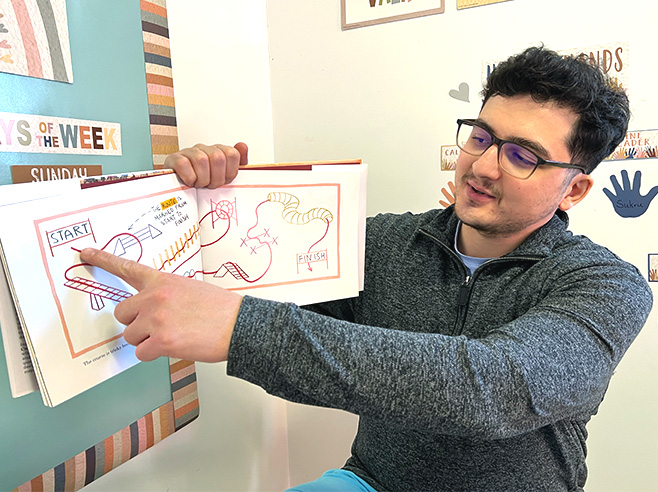 A man points to a pathway around landmarks in Julie Dillemuth’s book “Mapping My Day” as he holds up the book.