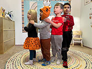 Four children stand in a line in the classroom holding onto the shoulders of the child in front.