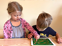 A girl plays the Gracie & Friends “Map Adventures” preschool spatial thinking app while a boy watches.