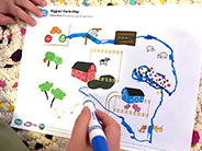 A boy draws a pathway with a marker between, over, around, and through landmarks on a printout of a farm map.