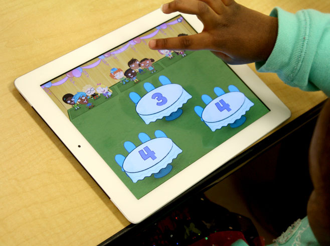 Close-up of a young child’s hand hovering over his iPad, where he is seating groups of characters at tables.