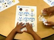 Café Lotto Match game materials are on a table, with children’s hands seen holding a game board and a set of cards.