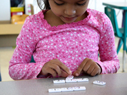 A girl in a pink shirt sits at a table and lines up dominoes with matching numbers.