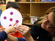 Close-up of a child’s hand pointing to a purple dot on the paper Dot Plate his teacher holds.