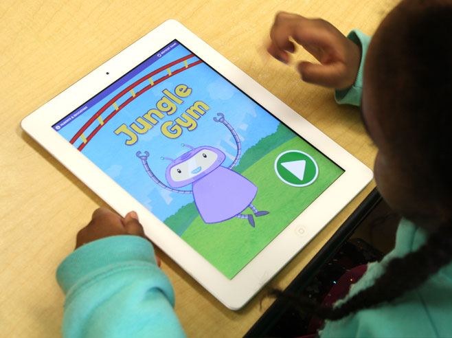 The home screen of the Gracie and Friends Jungle Gym app is seen on an iPad, with a young girl’s hands nearby.