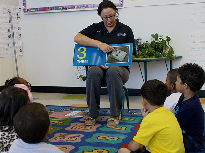 A teacher points to a page with 3 dots next to the number 3. A group of children sit on a rug and watch.