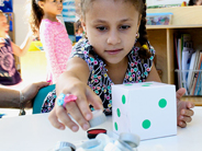A preschool girl with a large die in front of her reaches for bottle caps in a bowl.