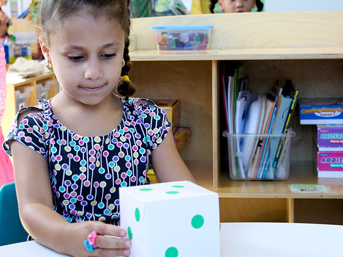 A girl holds and looks at a large die showing three green dots on top.
