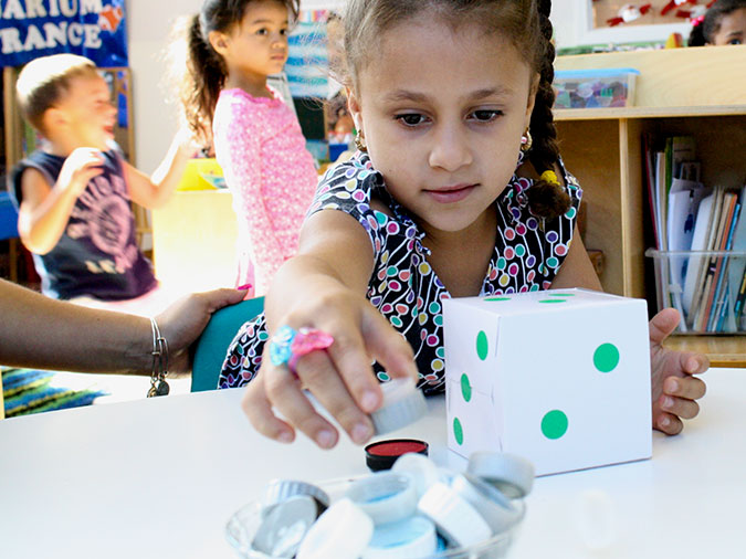 A preschool girl with a large die in front of her reaches for bottle caps in a bowl.