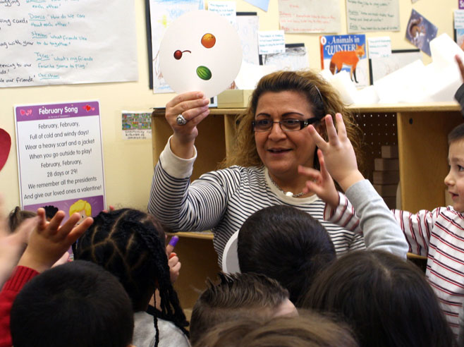 Preschoolers hold up three fingers as their teacher shows a round card with pictures of three fruits on it.