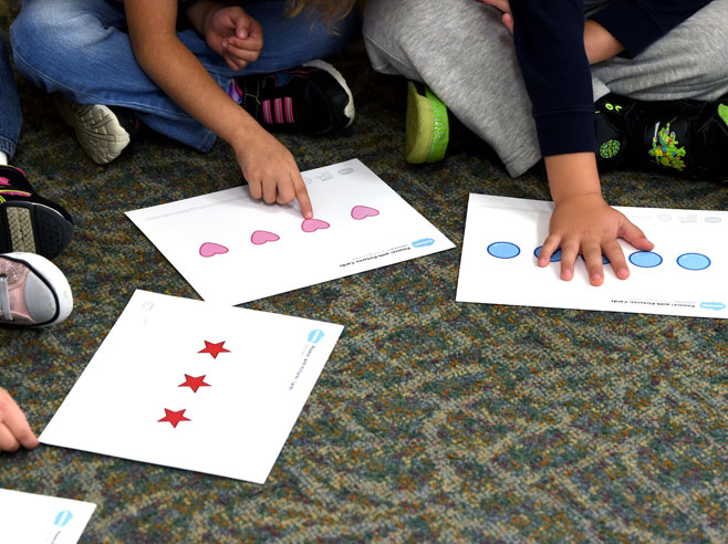Children seated on a carpet point to large white cards with several colorful objects.