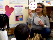 A teacher holds up a round card with four purple dots, while several preschool children watch.