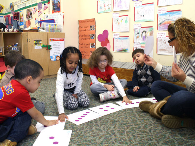 Children place their hands on cards on the carpet in front of them. A teacher holds up a round card and watches.