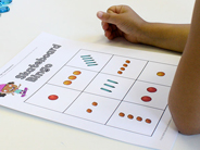 Close-up of a child resting her arms nest to a Skateboard Bingo board, a grid with one to five colorful objects in each square.