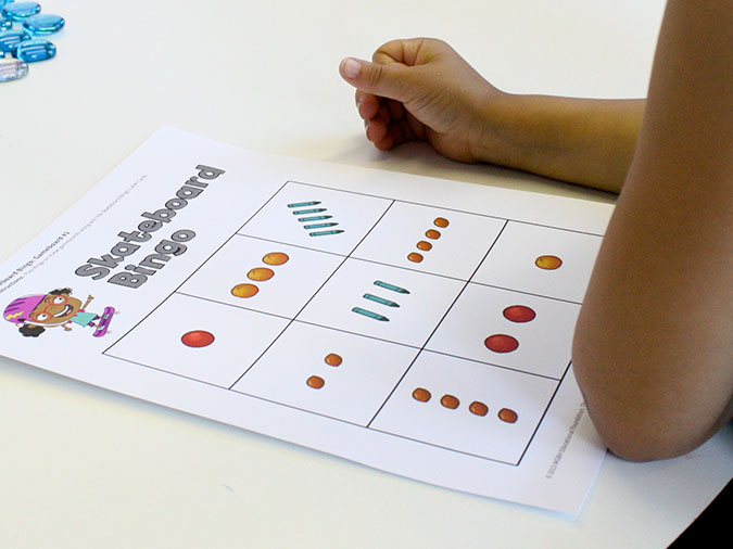 Close-up of a child resting her arms nest to a Skateboard Bingo board, a grid with one to five colorful objects in each square.