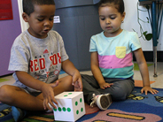 A young boy and girl sit on a carpet and look at two dots on a large die.