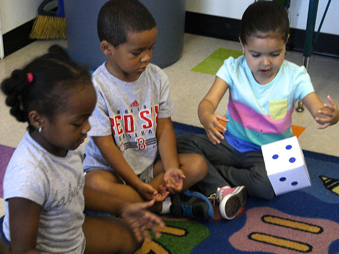 A girl sits on a carpet with her classmates and throws a large die.