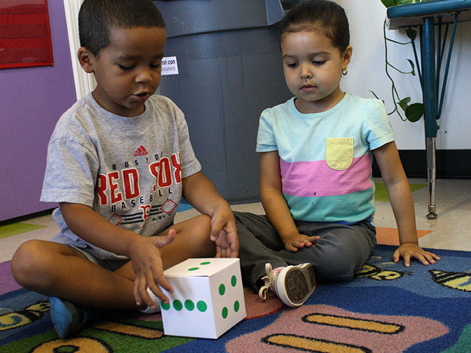 A young boy and girl sit on a carpet and look at two dots on a large die.