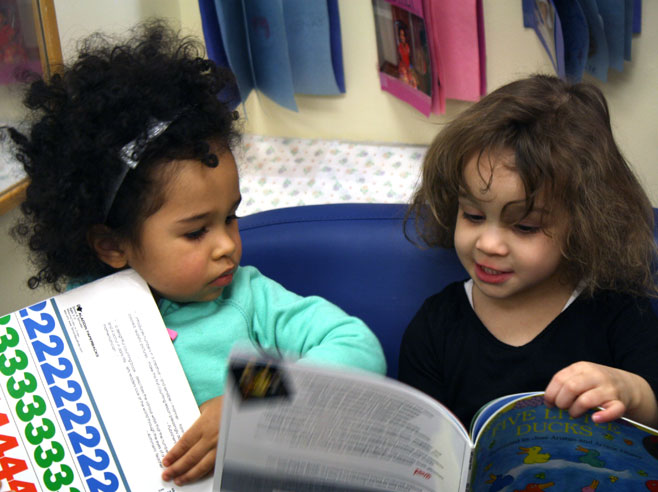 Two young girls sit on a small sofa and hold books. One girl shows a page of her book to the other girl.