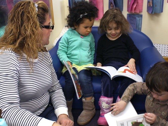 A preschool teacher sits near three children who are looking at books.