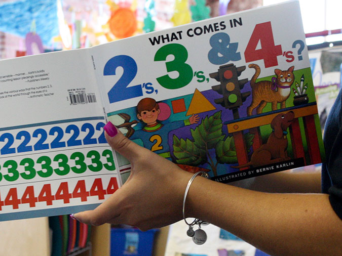 A teacher holds up the book What Comes In 2s, 3s & 4s?.