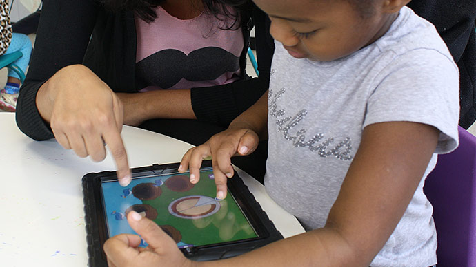 A preschool child in a gray shirt plays the Early Math with Gracie & Friends Photo Friends app and drags a piece of pie while a a teacher sits nearby and points out the birds’ nests where the pie should be placed.